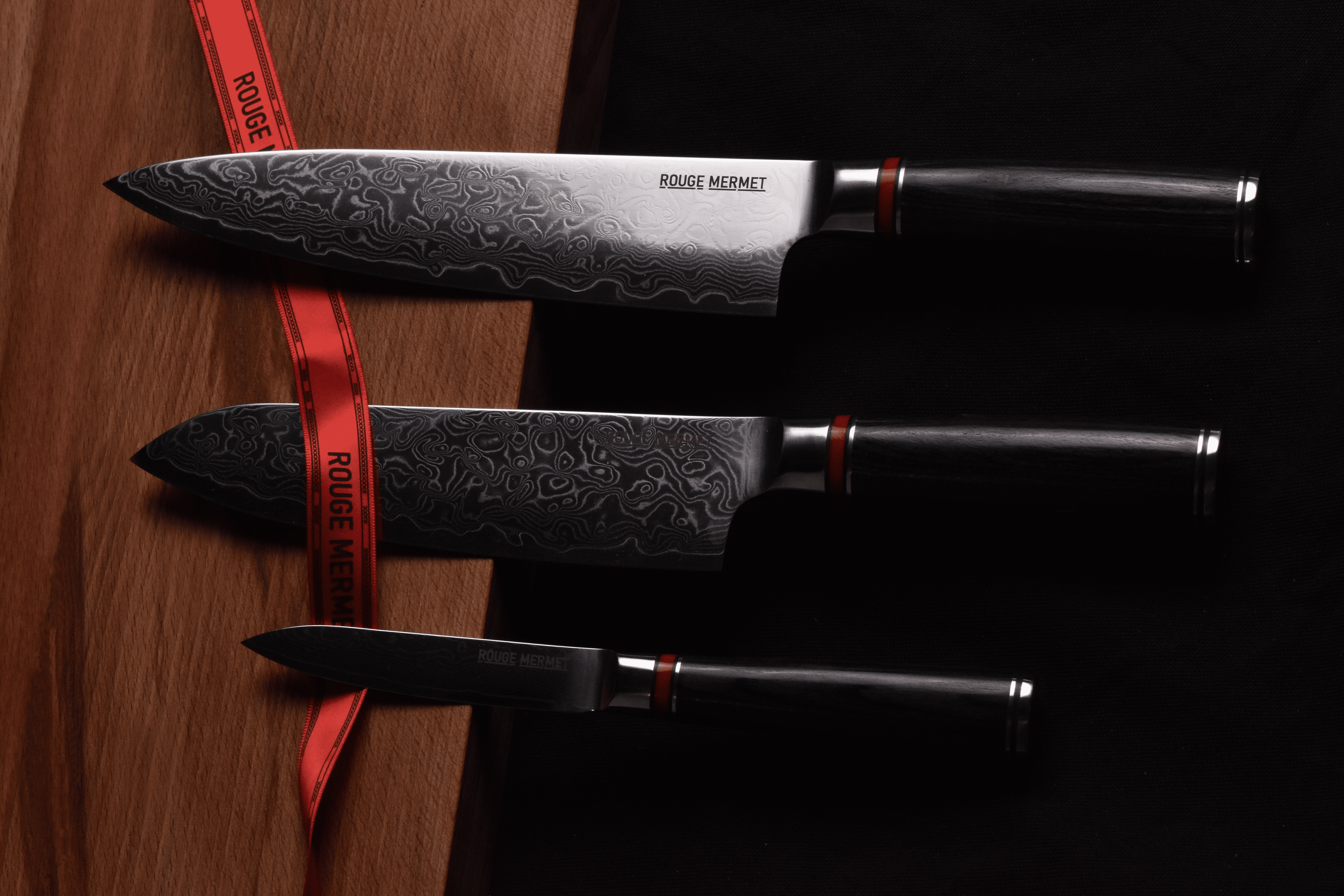3 couteaux de cuisine de haute qualité en acier VG10 et en damas. Ils sont de la marque ROUGE MERMET ont un manche en bois noir et un anneau rouge sur le manche qui est l'héritage de la marque. Ils sont posés sur une planche à découper en chêne massif et sont entourés d'un ruban rouge